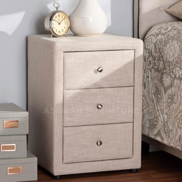 A beige fabric-upholstered nightstand with three drawers, topped with a clock and vase.