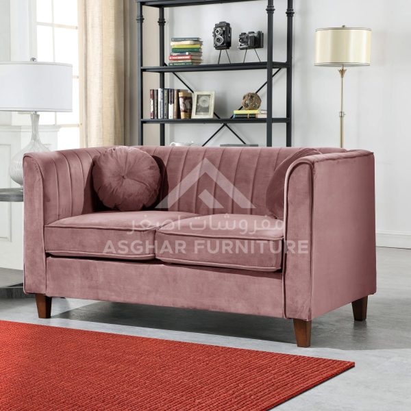 A modern two-seater sofa in a muted pink velvet with channel tufting on the back and a matching round cushion.