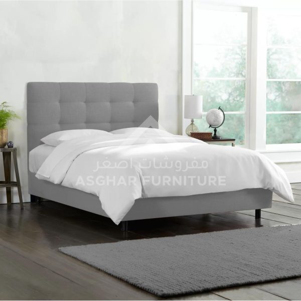 A simple gray tufted upholstered bed with a plain gray rug.