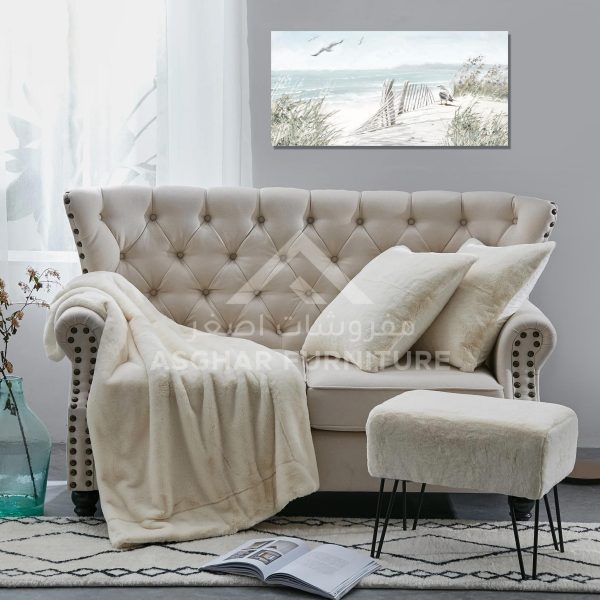 A two-seater tufted cream sofa with nailhead trim and rolled arms, with a matching fuzzy footstool with thin black legs.