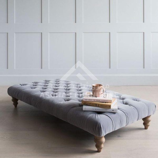 A low, tufted grey velvet ottoman with light, turned wood legs, serving as a coffee table with books and a mug on top.