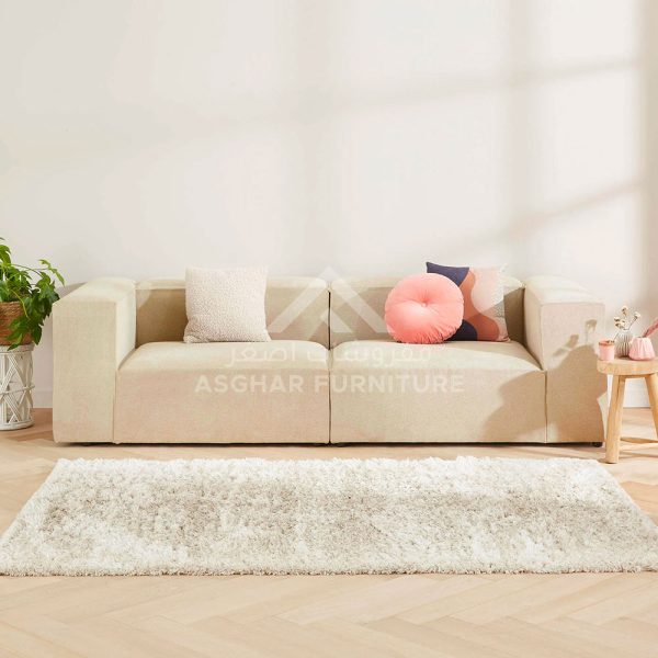 A light beige fabric modular sofa with decorative pillows, and a large fluffy white rug.