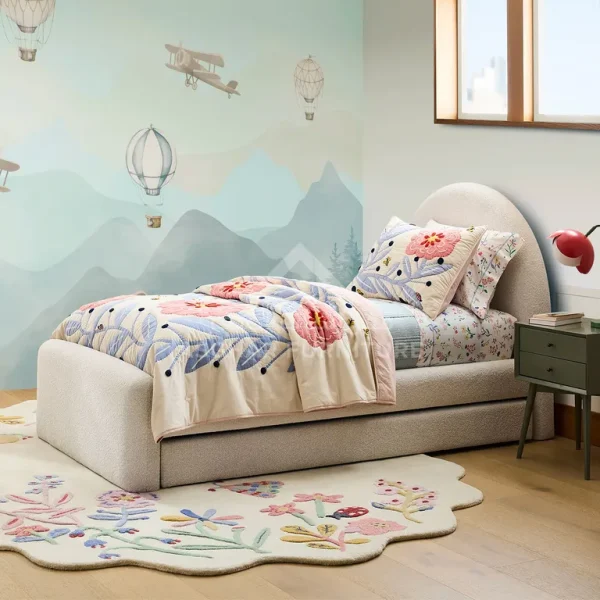 A whimsical child's bedroom features an upholstered bed with a floral comforter and a shaped rug, against a painted mural.
