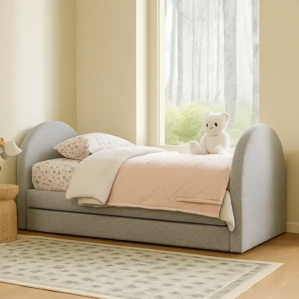 A cozy child's grey upholstered bed with a pull-out trundle, light bedding, and a white teddy bear.