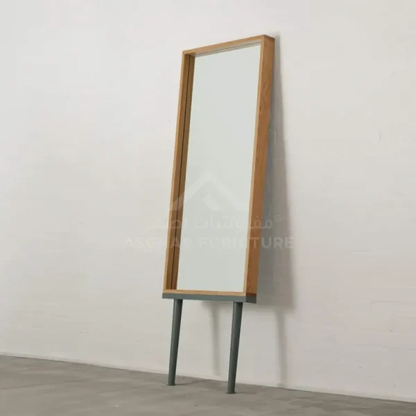 A contemporary rectangular mirror with a light wood frame, grey base, and two slender legs, leaning against a wall.