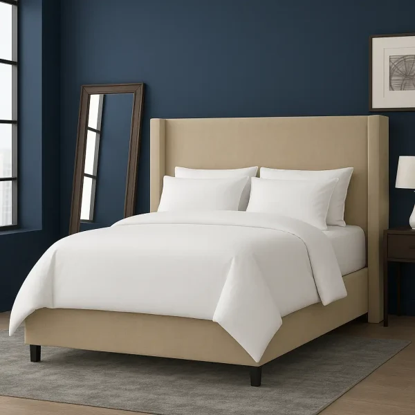 A neutral wingback bed with white bedding, a leaning mirror, and a nightstand with a lamp.