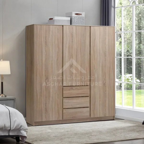 A modern three-door light wood wardrobe with three central drawers, standing next to a large window.
