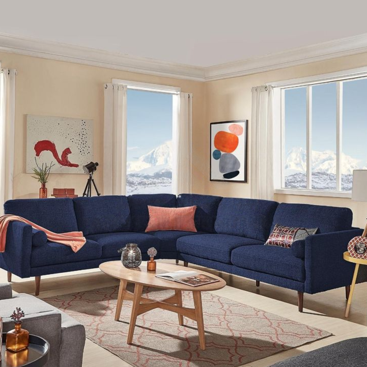 A large navy blue corner sectional sofa with mid-century style legs, a wooden coffee table, and windows showing a snowy mountain scene.