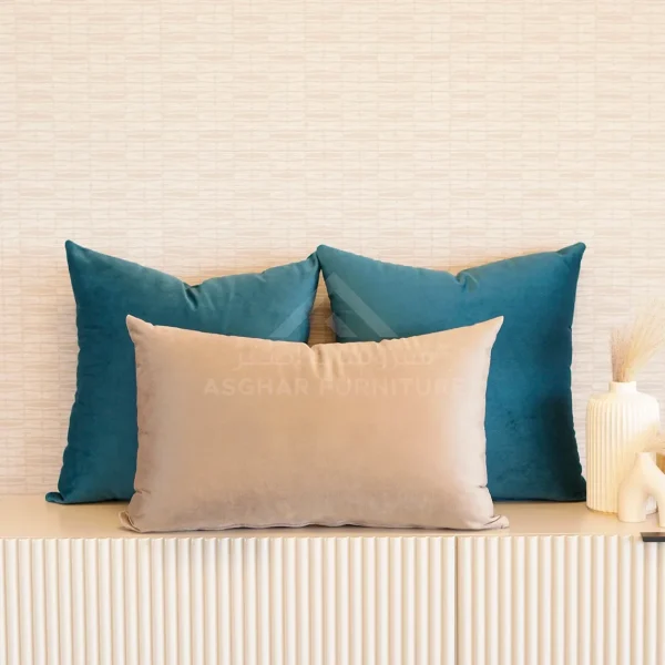 Two square dark teal cushions behind a rectangular beige cushion, arranged on a ridged console table.
