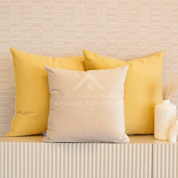 A soft, square cream-colored velvet cushion placed in front of two larger yellow cushions.