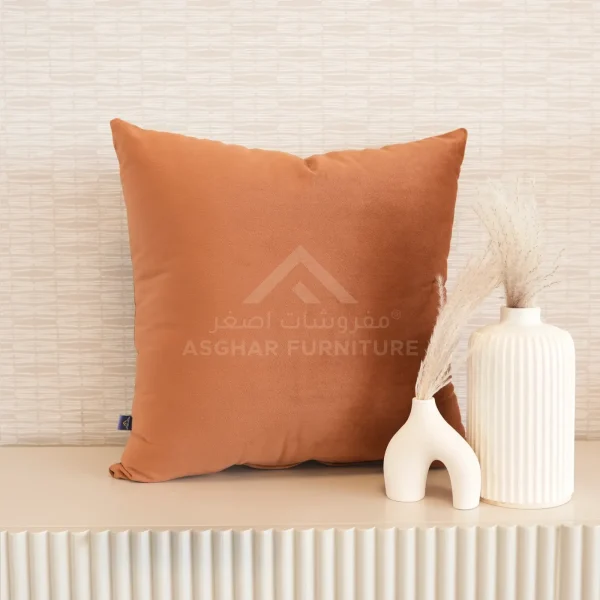 A soft, square terracotta-colored velvet cushion next to two modern white vases with dried grasses.