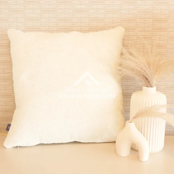 A large, square cream-colored fuzzy cushion next to two modern white vases with dried grasses.
