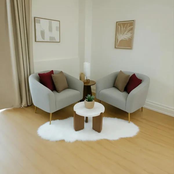 A pair of light gray armchairs with gold legs, red and brown pillows, and a small, round coffee table on a furry white rug.