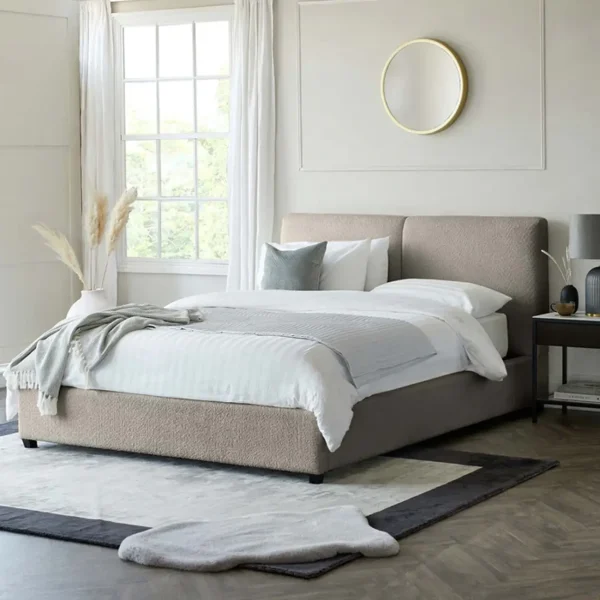 A beige upholstered bed with white bedding, gray accents, a large window, and a round gold mirror.