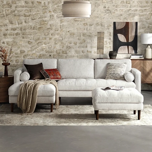 A beige tufted sectional sofa with an ottoman and various pillows sits in a room with a stone wall.
